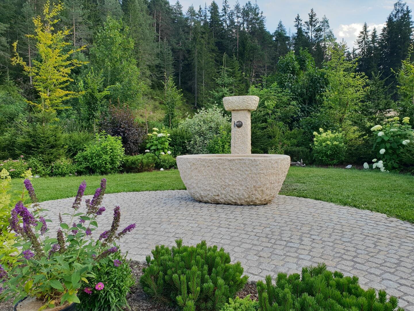Ein steinerner Wasserbrunnen steht auf einem runden Kopfsteinpflaster, umgeben von grünen Sträuchern, bunten Blumen und dichten Bäumen in einer üppigen Gartenlandschaft.