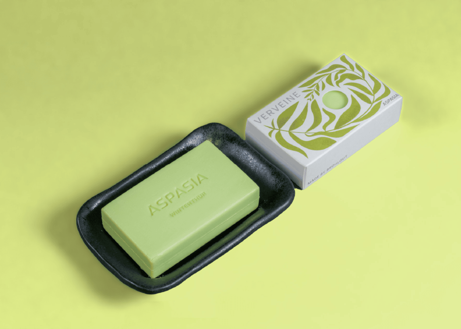 A green bar of Aspasia soap on a black dish lies next to the white and green Verveine box, both on a light green background.
