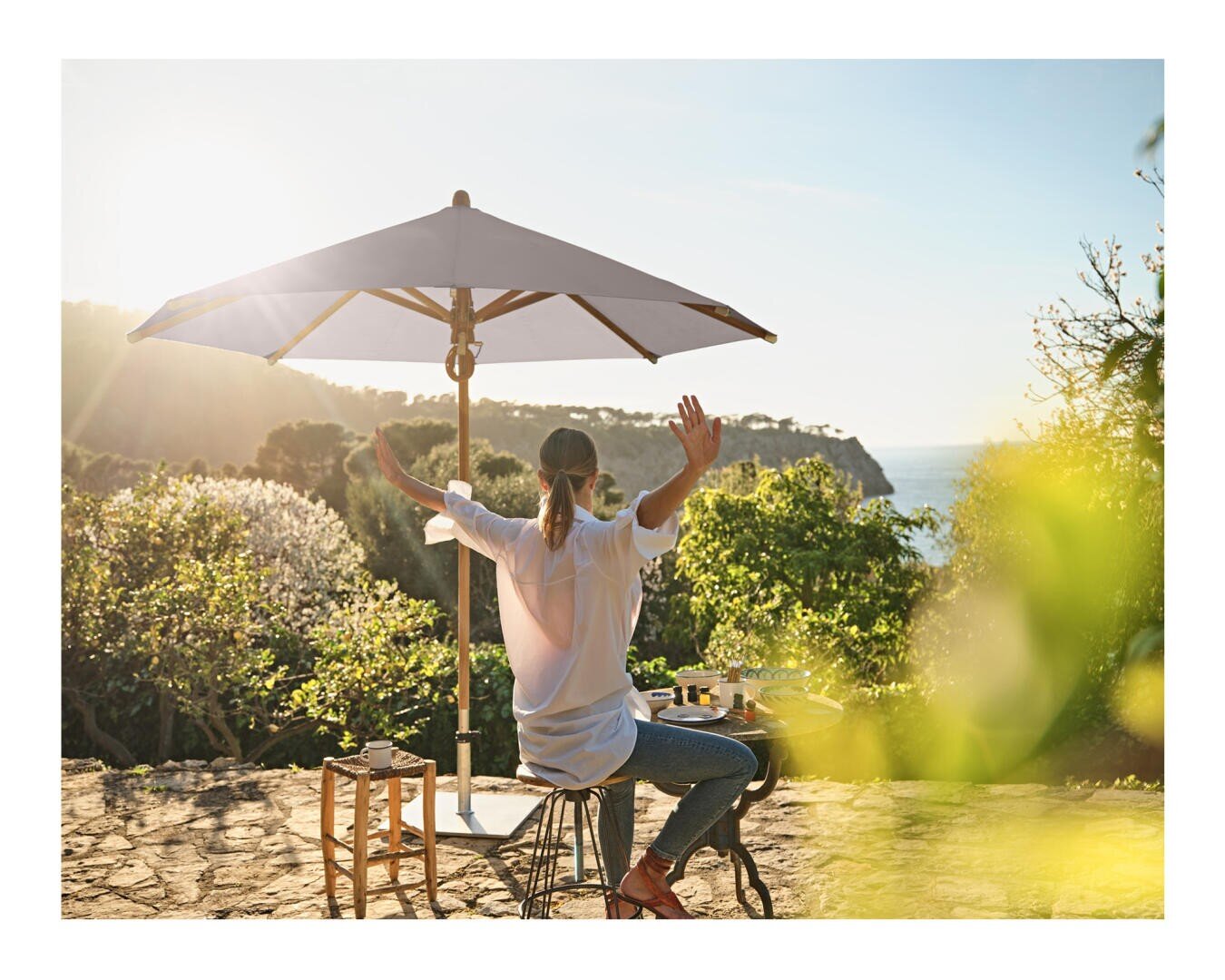 Eine Person sitzt an einem Tisch im Freien unter einem großen Sonnenschirm, die Arme ausgestreckt, und genießt die Aussicht auf üppiges Grün und das Meer im Hintergrund an einem sonnigen Tag.