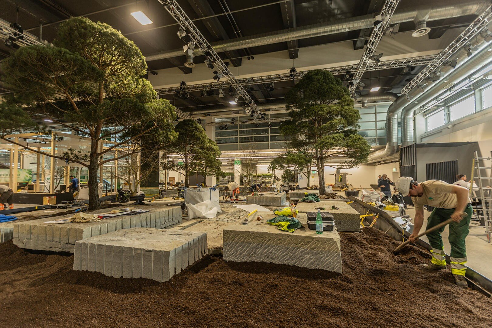 In einer geräumigen Halle mit Industriebeleuchtung und freiliegenden Rohren schichten Arbeiter Erde und Steinplatten um mehrere große Bäume herum auf. Baumaterialien und -geräte sind überall in der Szene verstreut.