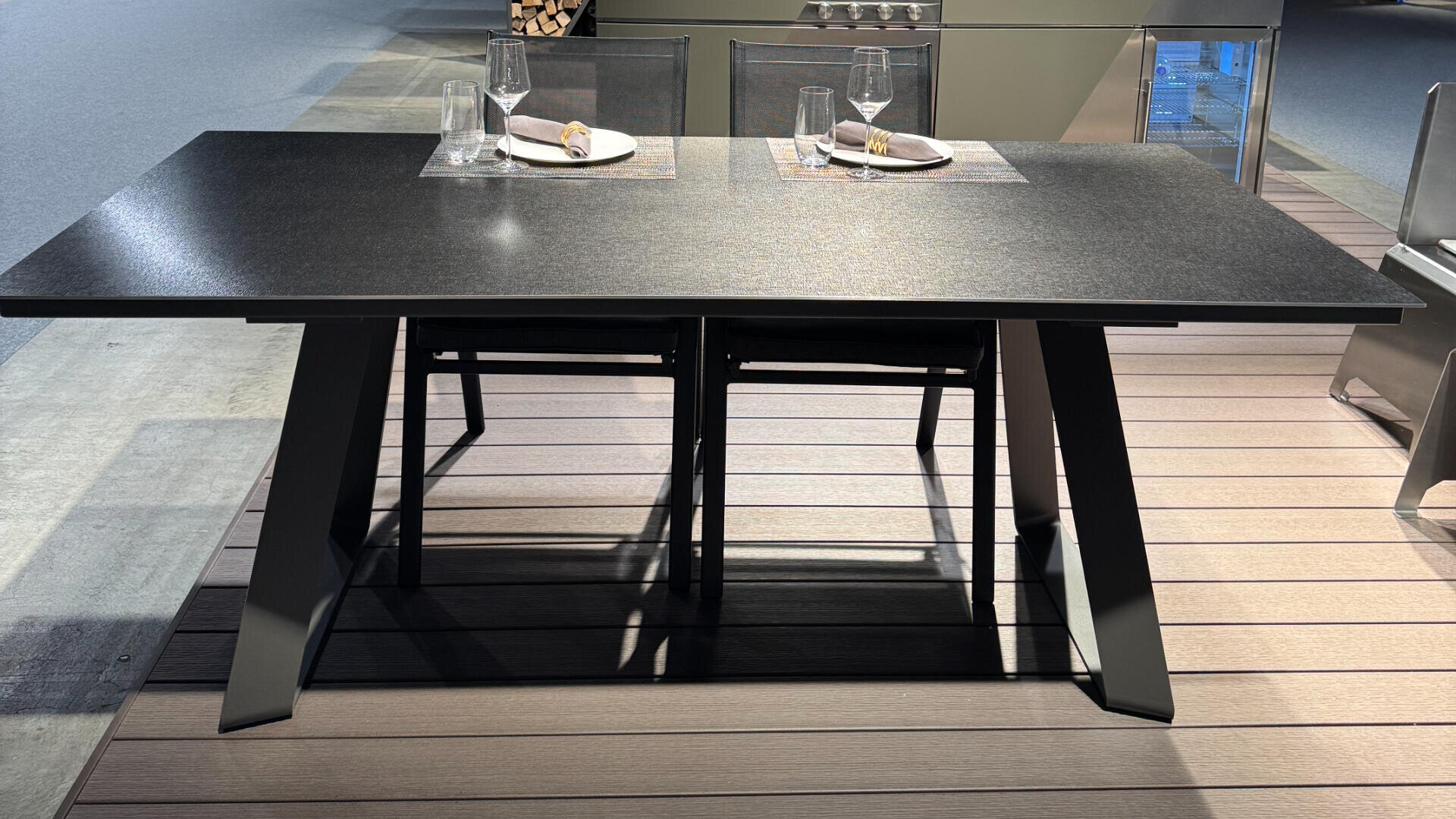 Ein moderner rechteckiger Esstisch mit dunkler Oberfläche und zwei Gedecken mit je einer Serviette, einem Teller, einem Weinglas und einem Wasserglas auf einem Deck mit zwei Stühlen.