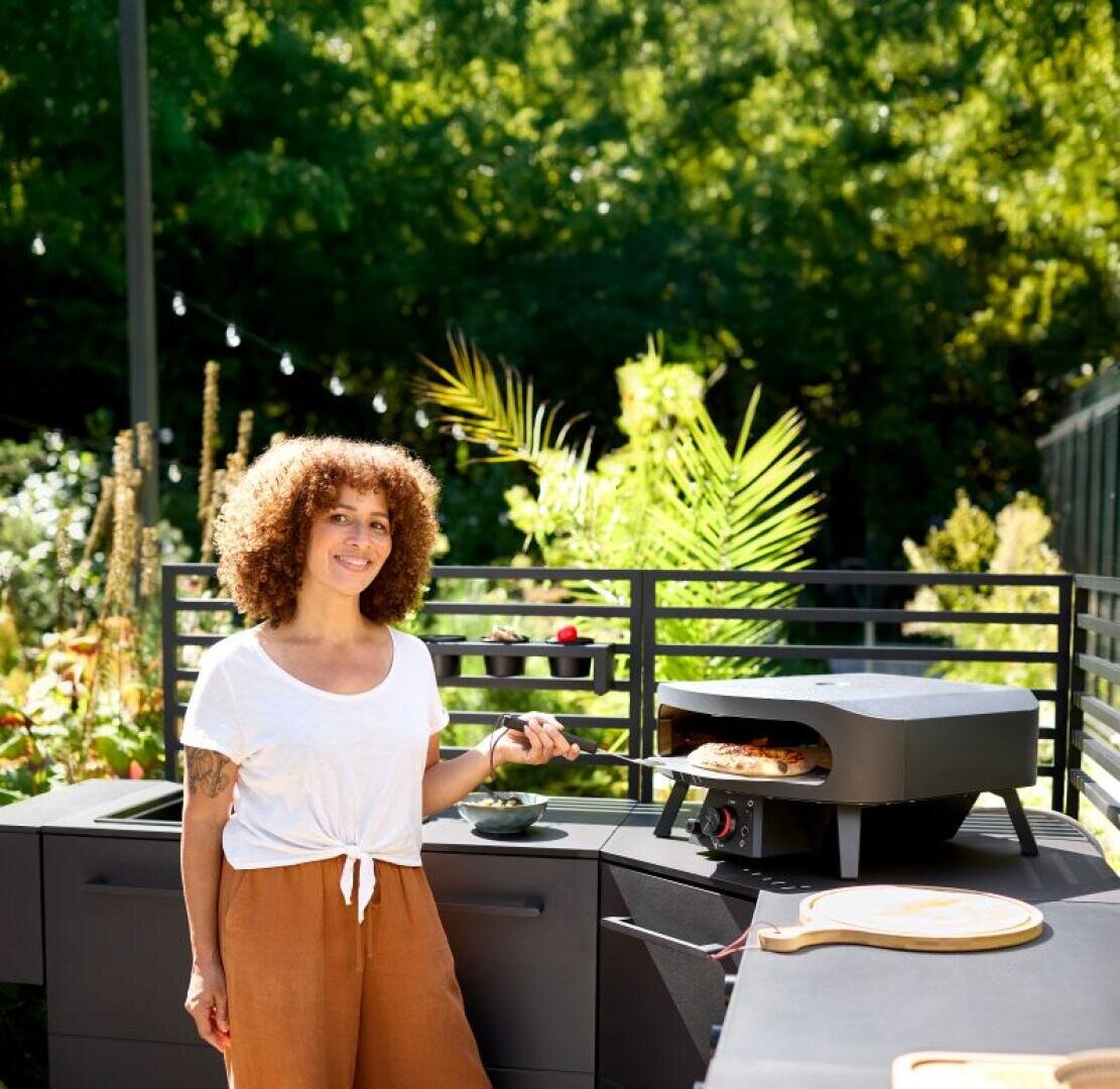 A woman with curly hair, wearing a white shirt and brown trousers, smiles as she stands outdoors next to a black pizza oven with a pizza inside. Green plants and trees can be seen in the sunny background.