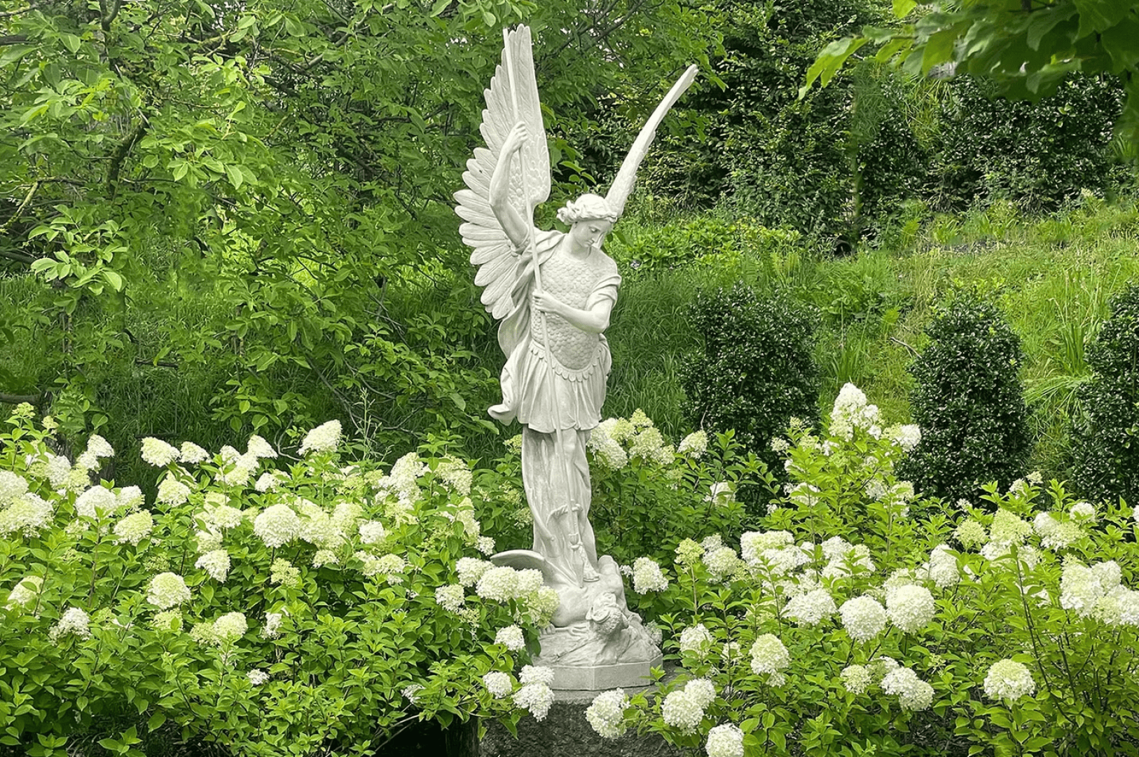 A white stone statue of an angel with wings stands among blooming white flowers and lush green bushes in a garden. The angel holds a sword and looks off to the side. The background is filled with greenery.