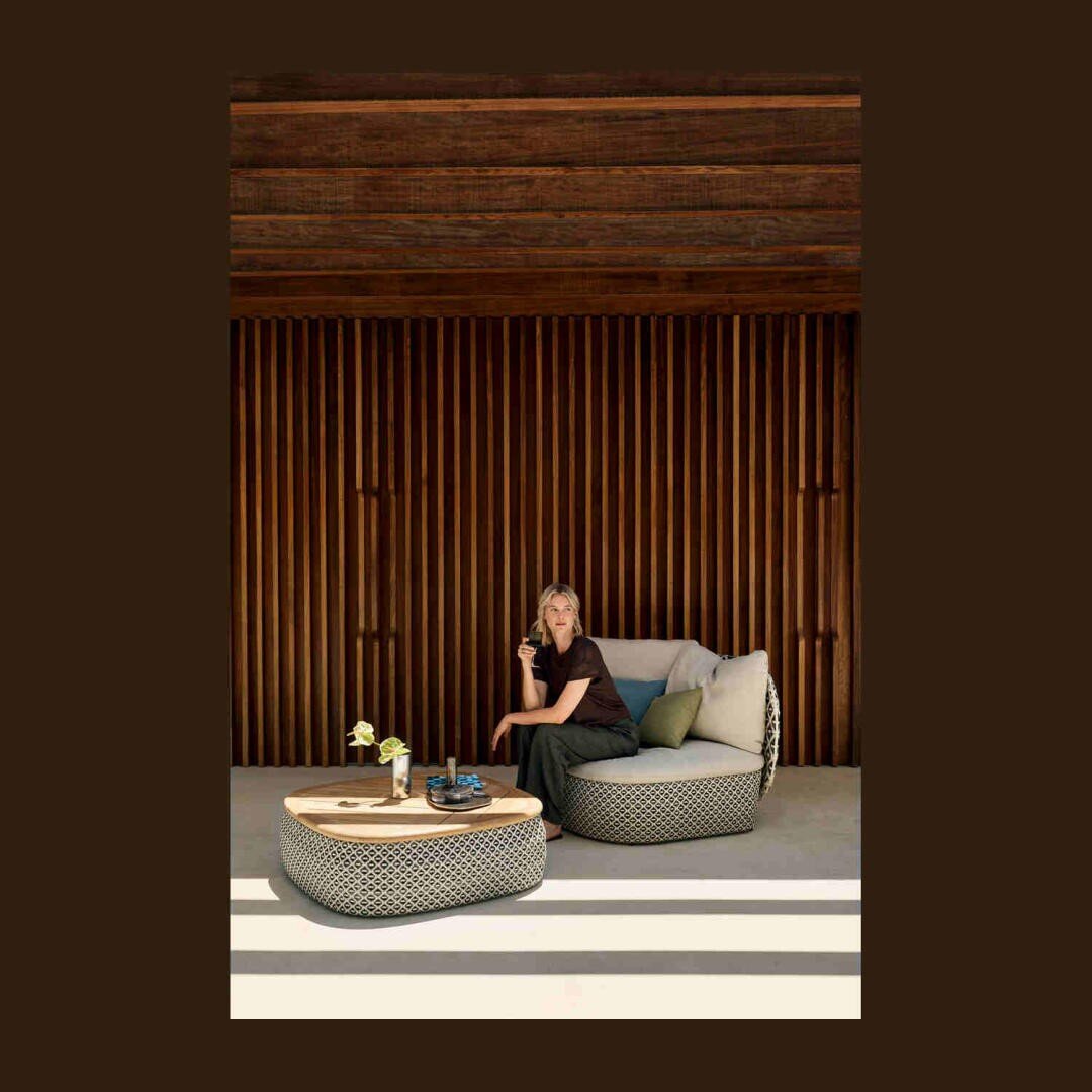 Eine Person sitzt auf einem modernen Outdoor-Sofa neben einem passenden Couchtisch mit Pflanzen und Getränken vor einer Wand mit Holzlatten und einem Dach, wobei das Sonnenlicht Schatten auf den Boden wirft.