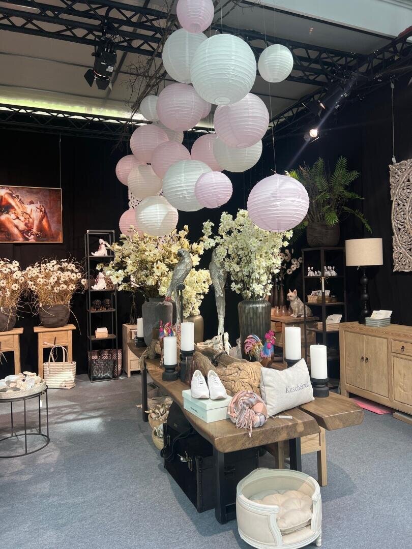 Eine dekorative Auslage zeigt einen Holztisch, über dem weiße und rosafarbene Papierlaternen hängen, umgeben von Vasen mit weißen Blumen, Kerzen, Kissen, Schuhen und verschiedenen Einrichtungsgegenständen in einem gemütlichen Innenbereich.