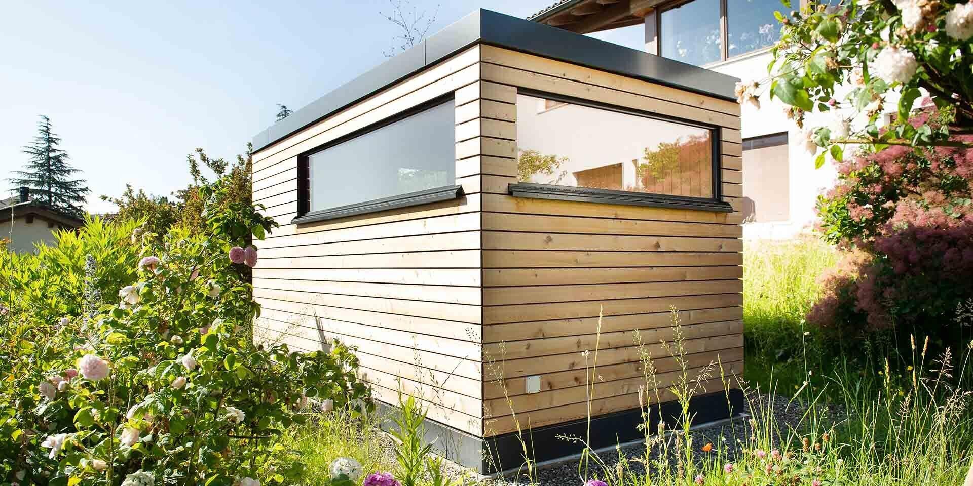Ein kleiner, moderner Holzschuppen mit horizontaler Verkleidung und einem großen rechteckigen Fenster steht in einem üppigen, grünen Garten, der von verschiedenen Pflanzen und Blumen umgeben ist.