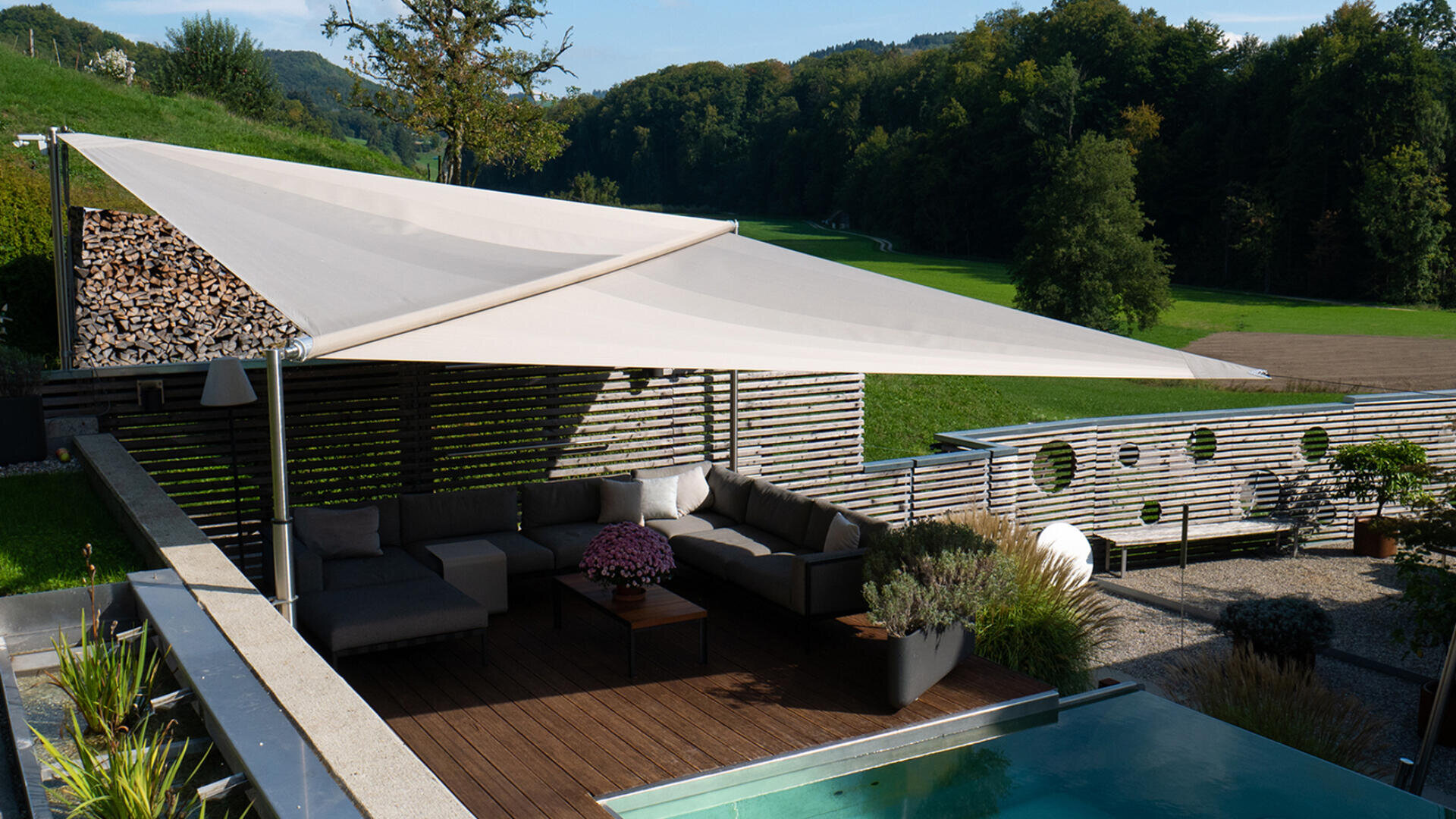 Moderner Sitzbereich im Freien mit einem großen dreieckigen Sonnensegel, Sektionssofas, Pflanzen und einem kleinen Pool auf einer grünen Rasenfläche mit Bäumen und Hügeln im Hintergrund.