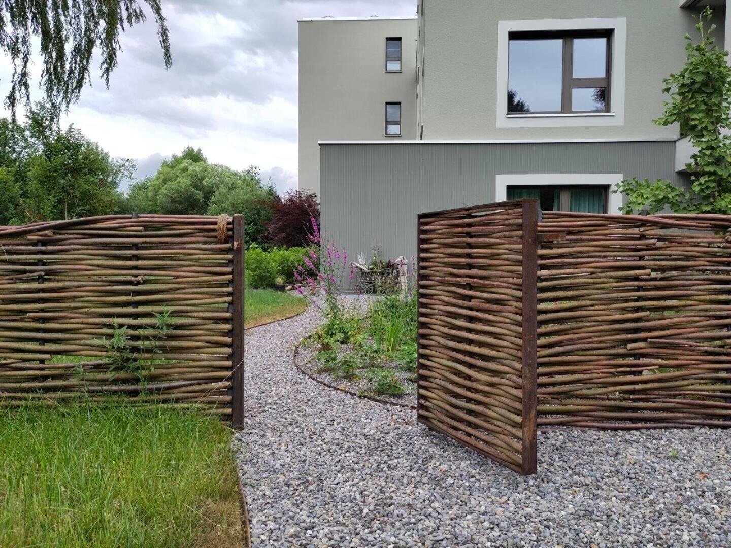A woven wooden gate stands open and leads to a gravel path with purple flowers and green plants in front of a modern gray building with large windows under a cloudy sky.