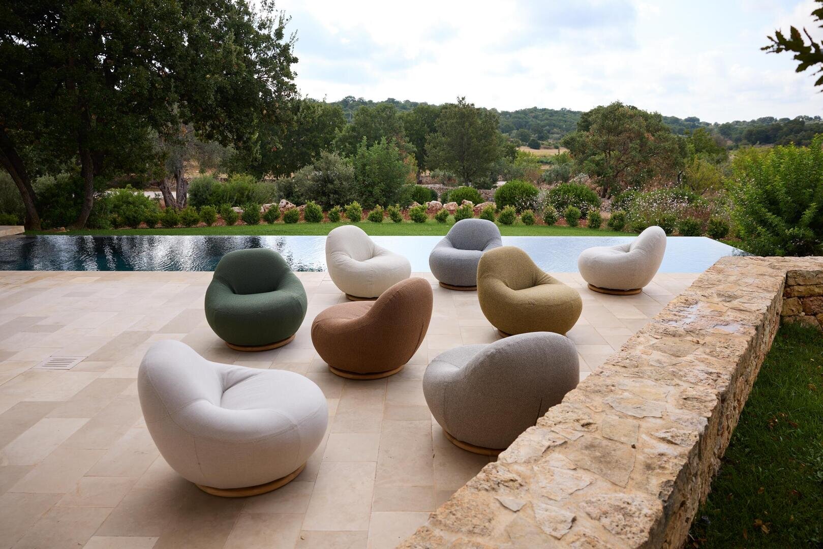 Mehrere moderne, runde Lounge-Sessel in neutralen und pastellfarbenen Tönen stehen auf einer Steinterrasse neben einem spiegelnden Pool, mit üppigem Grün und Bäumen im Hintergrund.