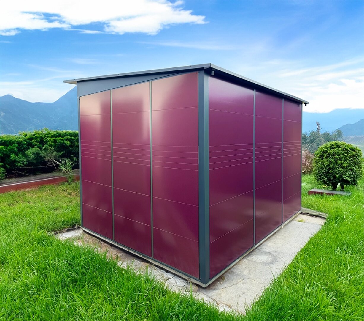 A modern, maroon metal shed with a flat roof stands on a concrete slab, surrounded by green grass, bushes and mountains in the background under a blue sky with scattered clouds.