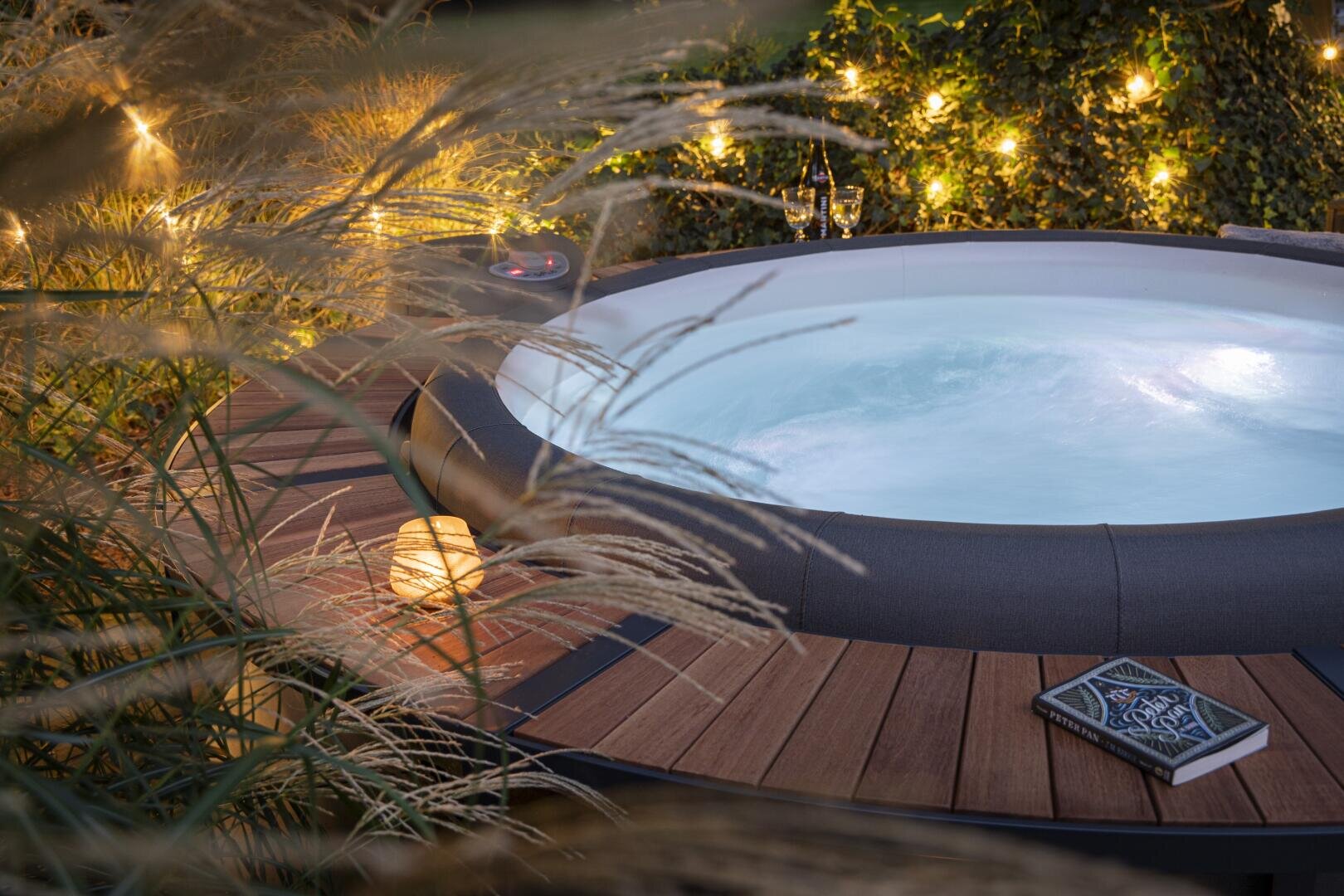 A bubbling hot tub on a wooden deck is surrounded by tall grass and warm string lights. Nearby, a closed book and a glowing lantern create a cozy, tranquil outdoor atmosphere in the evening.