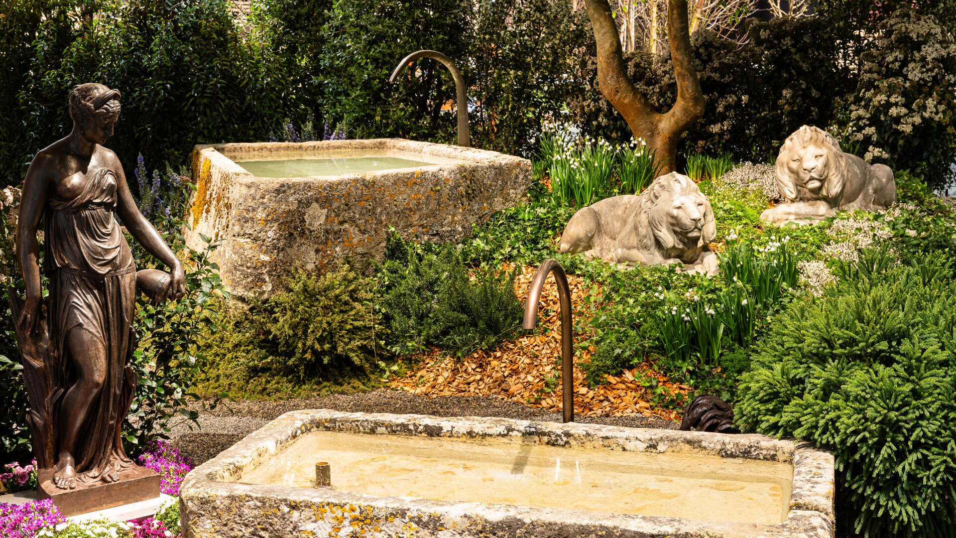 Eine Gartenszene mit zwei Steinbrunnen, üppigen grünen Pflanzen, einer Bronzestatue einer Frau und zwei steinernen Löwenskulpturen, die im Gras neben einem Baum ruhen. Das Sonnenlicht erhellt die heitere, dekorative Umgebung.