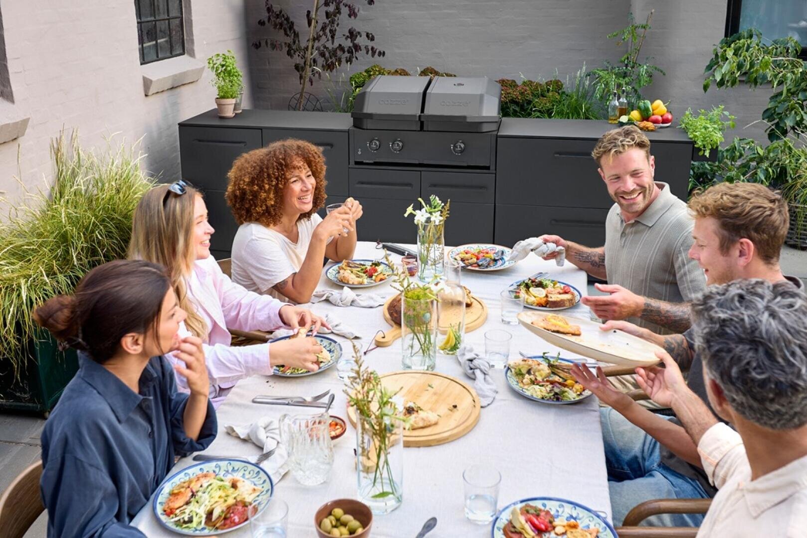 A group of six people are sitting at an outdoor table enjoying a meal together, smiling and laughing. There are plates of food, drinks and empty dishes on the table. A barbecue and plants can be seen in the background.