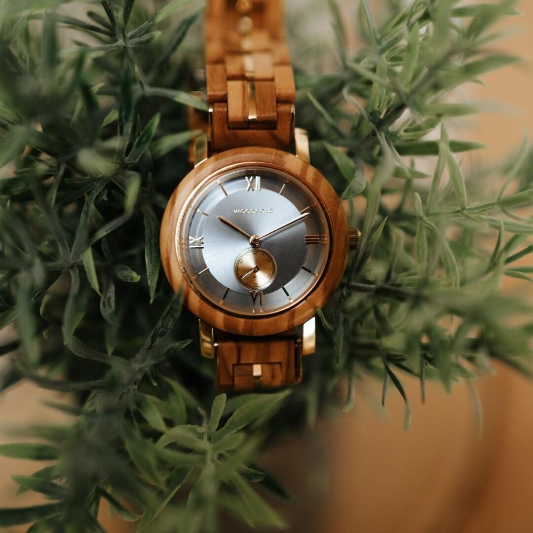 Eine hölzerne Armbanduhr mit silbernem Ziffernblatt und römischen Ziffern liegt inmitten grüner Blätterzweige.