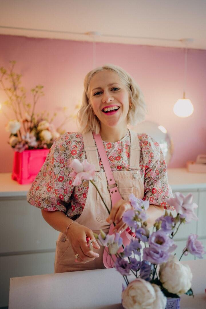 Eine lächelnde Frau mit kurzen blonden Haaren, die eine geblümte Bluse und einen hellrosa Overall trägt, arrangiert lila und weiße Blumen in einem hellen Raum mit rosa Wänden und sanfter Beleuchtung.