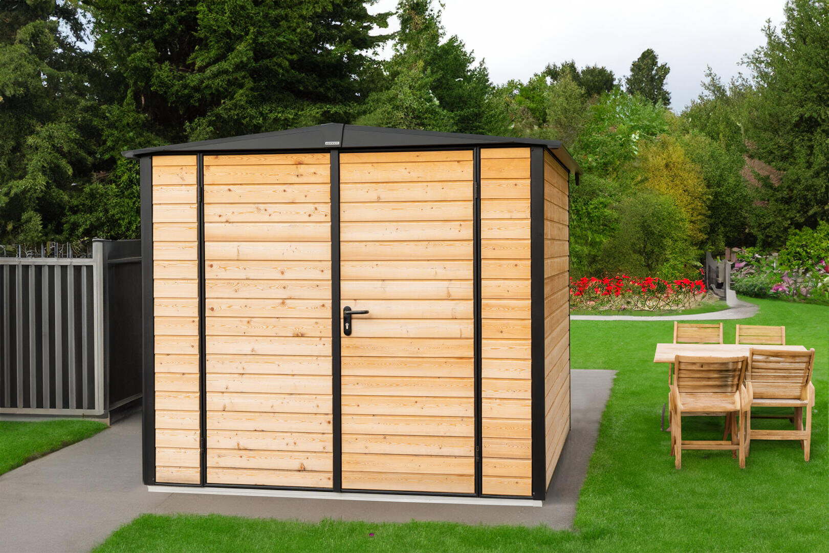 A wooden garden shed with a black frame and a locked door stands on a paved area next to a lawn with a wooden dining table and chairs; in the background are trees and colorful flowerbeds.