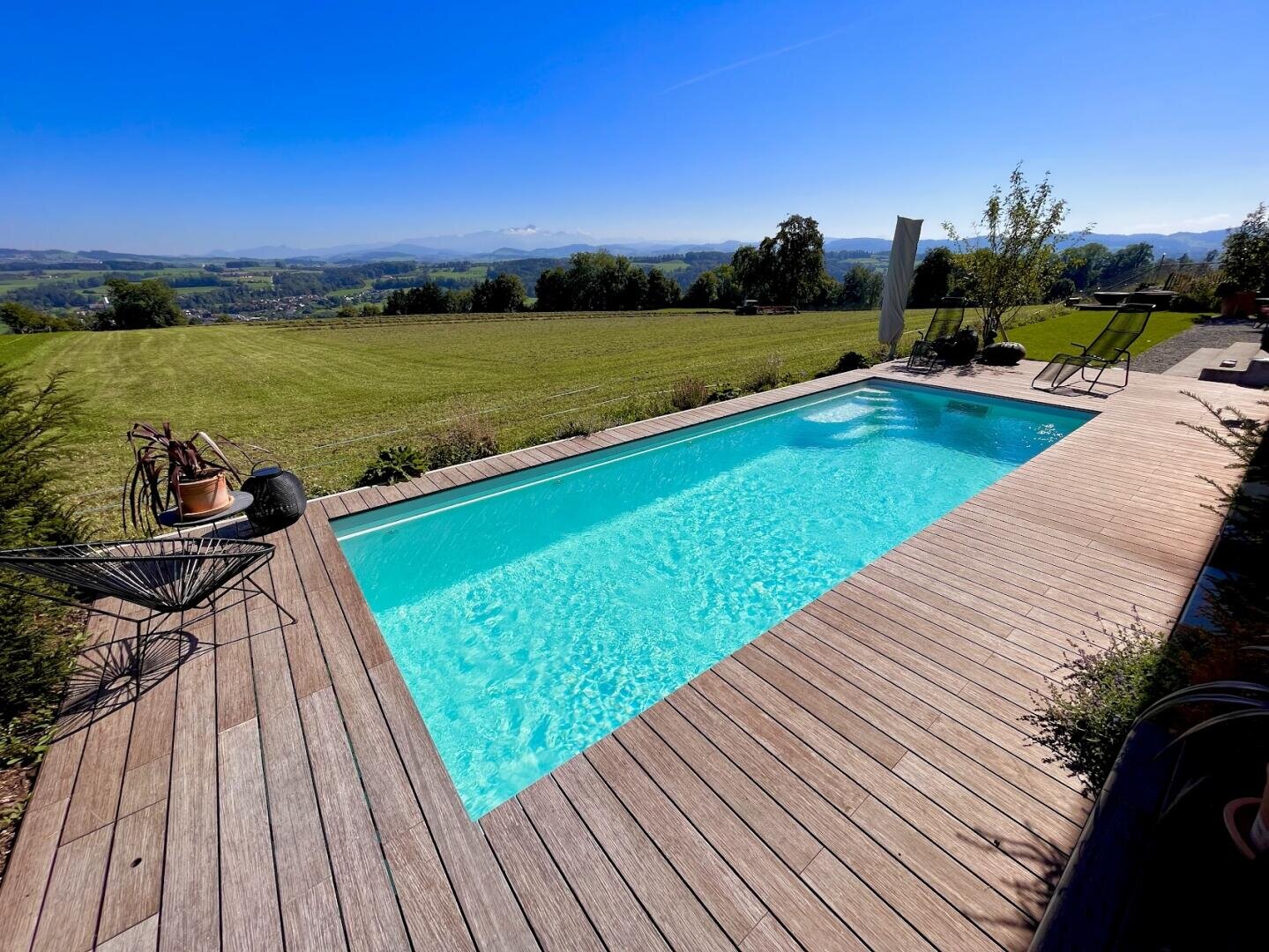 Ein rechteckiger Außenpool mit klarem, blauem Wasser ist von einer Holzterrasse umgeben, die einen Blick auf ein weites, grünes Feld und ferne Hügel unter einem blauen Himmel bietet. Liegestühle und Pflanzen schmücken die Terrasse.