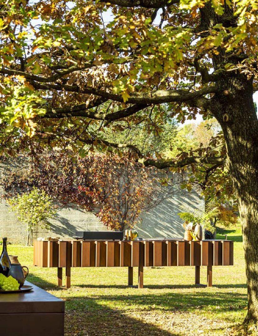 Moderne Outdoor-Möbel stehen auf einem sonnenbeschienenen Rasen unter einem großen Baum mit grünen und vergilbten Blättern und schaffen eine entspannte, natürliche Atmosphäre. Auf den Tischen im Hintergrund sind Dekorationsartikel und Getränke angeordnet.