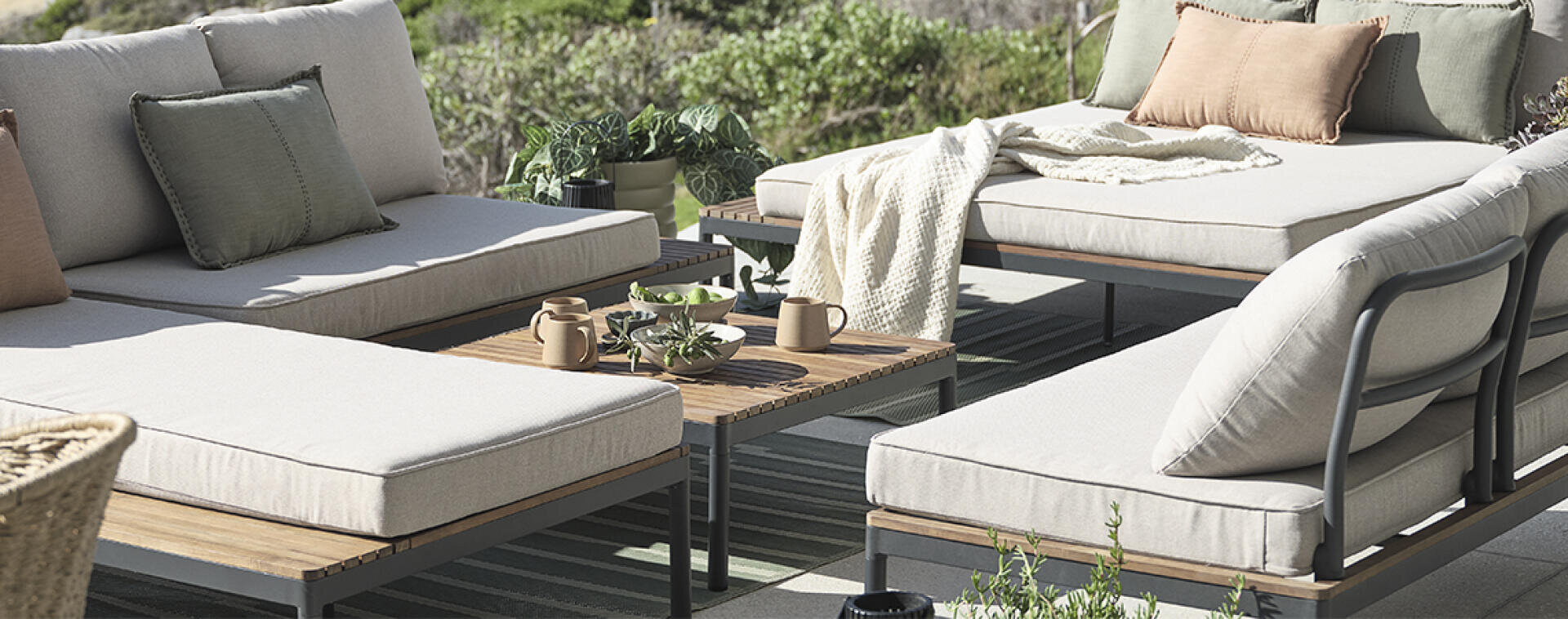 Terrasse im Freien mit beigefarbenen gepolsterten Loungesesseln, hölzernen Couchtischen, Topfpflanzen und neutralfarbenen Kissen und Decken auf einem gestreiften Teppich mit Grünzeug im Hintergrund.
