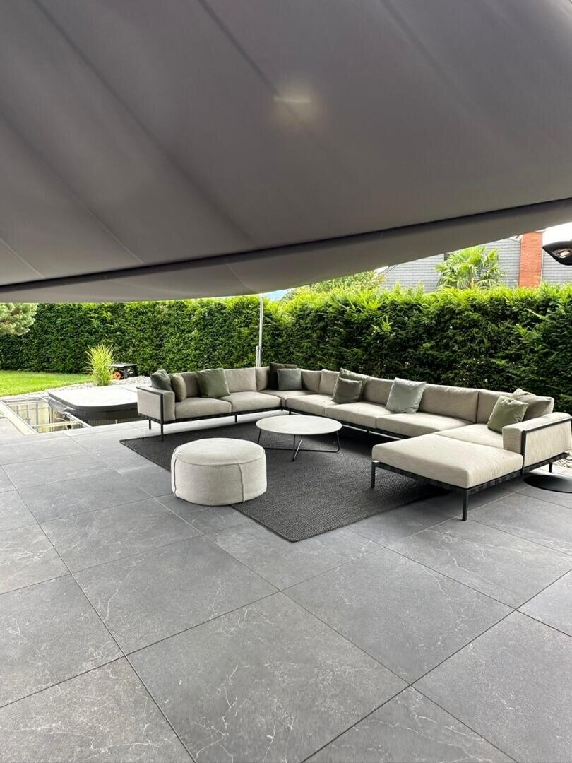 Moderne Außenterrasse mit großem grauen Sektionssofa, runder Ottomane und zwei runden Couchtischen auf einem dunklen Teppich. Umgeben von Grün und teilweise durch eine schlanke überhängenden Baldachin beschattet. Bodenbelag aus Steinfliesen im ganzen Haus.