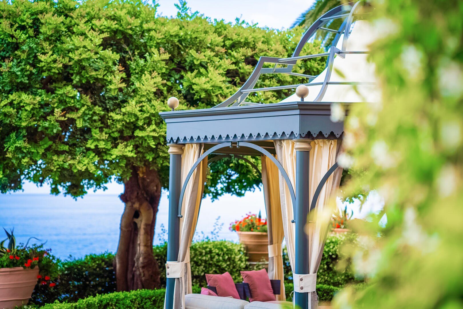 Elegant pavilion with metal columns and decorative roof, surrounded by lush greenery and potted plants. The sea and blue sky can be seen in the background. Partially upholstered seating can be seen under the gazebo.