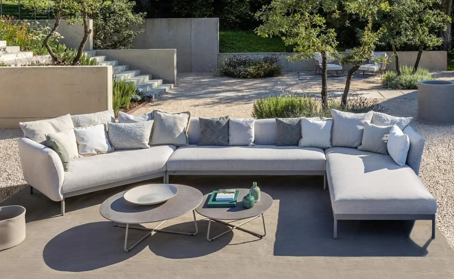 Eine moderne Außenterrasse mit einem hellgrauen L-förmigen Sektionssofa, zahlreichen Kissen, einem Couchtisch mit Büchern, Vasen und Schalen, alles auf einer Terrasse, umgeben von Kies, Bäumen und Grünpflanzen.