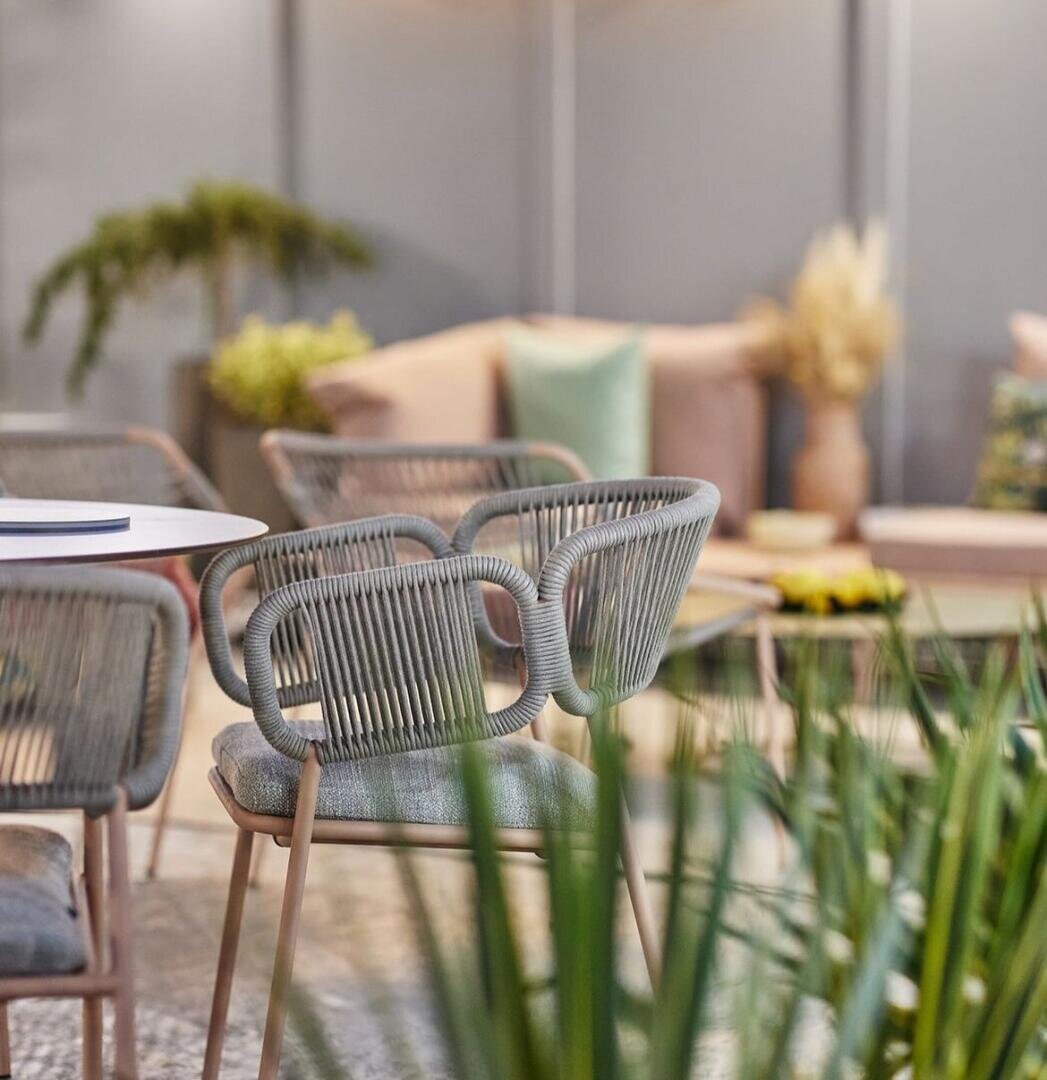 Moderne Sitzecke im Innenbereich mit grauen geflochtenen Stühlen um einen weißen Tisch. Im Vordergrund stehen Grünpflanzen, und im Hintergrund ist ein beiges Sofa mit pastellfarbenen Kissen und Pampasgrasdekor zu sehen. Die Beleuchtung ist sanft.