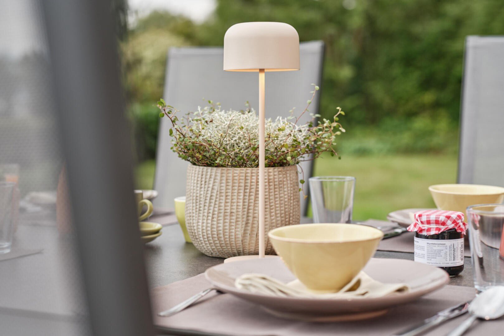Nahaufnahme eines Esstisches im Freien mit gelben Schalen, Tellern, einem geflochtenen Blumentopf mit Grünzeug, einer modernen weißen Lampe, Gläsern und kleinen Marmeladengläsern. Im unscharfen Hintergrund ist Grünzeug zu sehen.