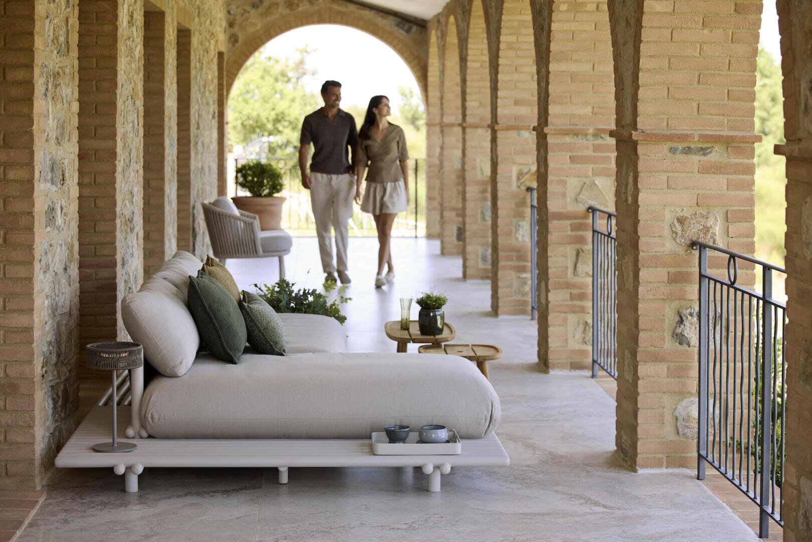 Ein Paar spaziert über eine überdachte Steinterrasse mit gewölbten Säulen. Im Vordergrund stehen moderne gepolsterte Loungesessel, kleine Tische und Topfpflanzen, die eine entspannte Atmosphäre im Freien schaffen.