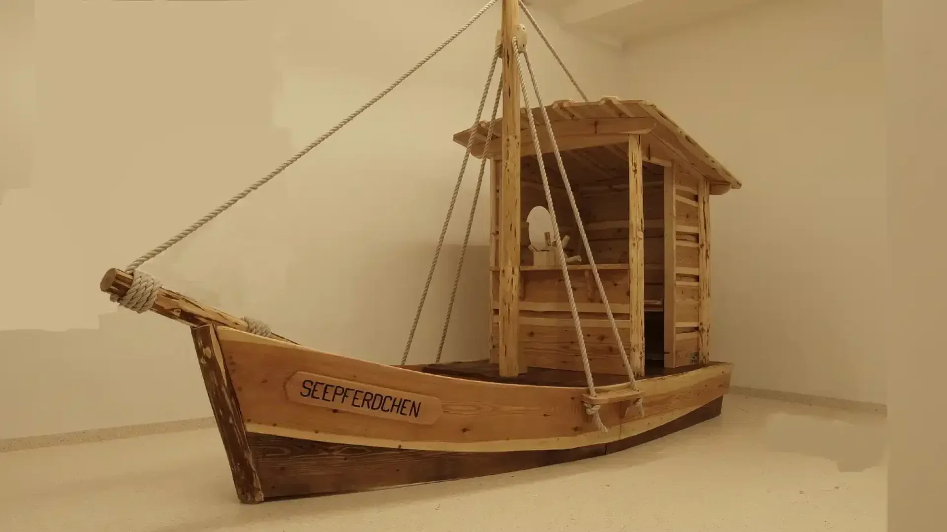 Ein hölzernes Spielboot mit einer kleinen Kajüte und einem Mast steht drinnen auf einem glatten, hellen Boden. An der Seite des Bootes steht das Wort SEEPFERDCHEN geschrieben.