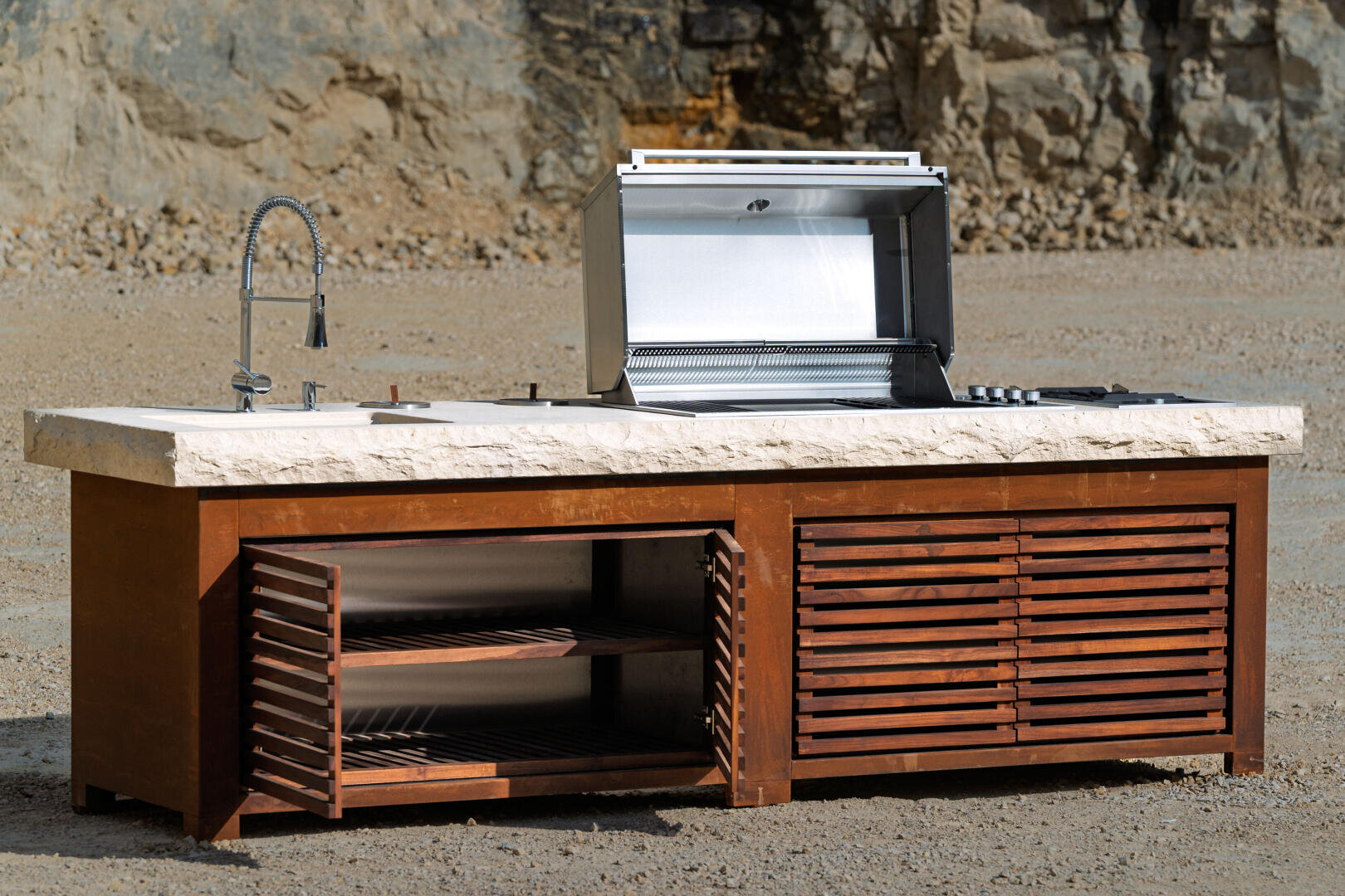 A modern outdoor kitchen unit with a built-in grill, open stainless steel lid, sink with a curved faucet, and wooden slatted cabinets, set on a gravel surface with a rocky background.