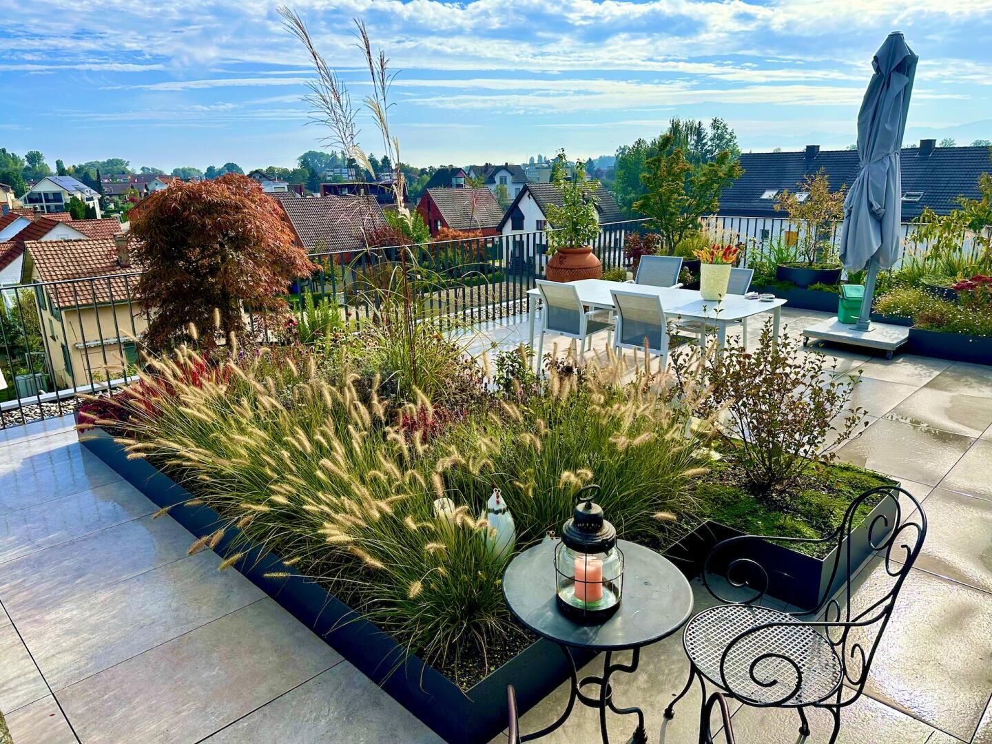 Eine Dachterrasse mit modernen Gartenmöbeln, üppigen Pflanzgefäßen mit Gräsern und Sträuchern, einem kleinen runden Tisch mit einer Laterne und einem Blick auf Dächer und Bäume unter einem strahlend blauen Himmel.
