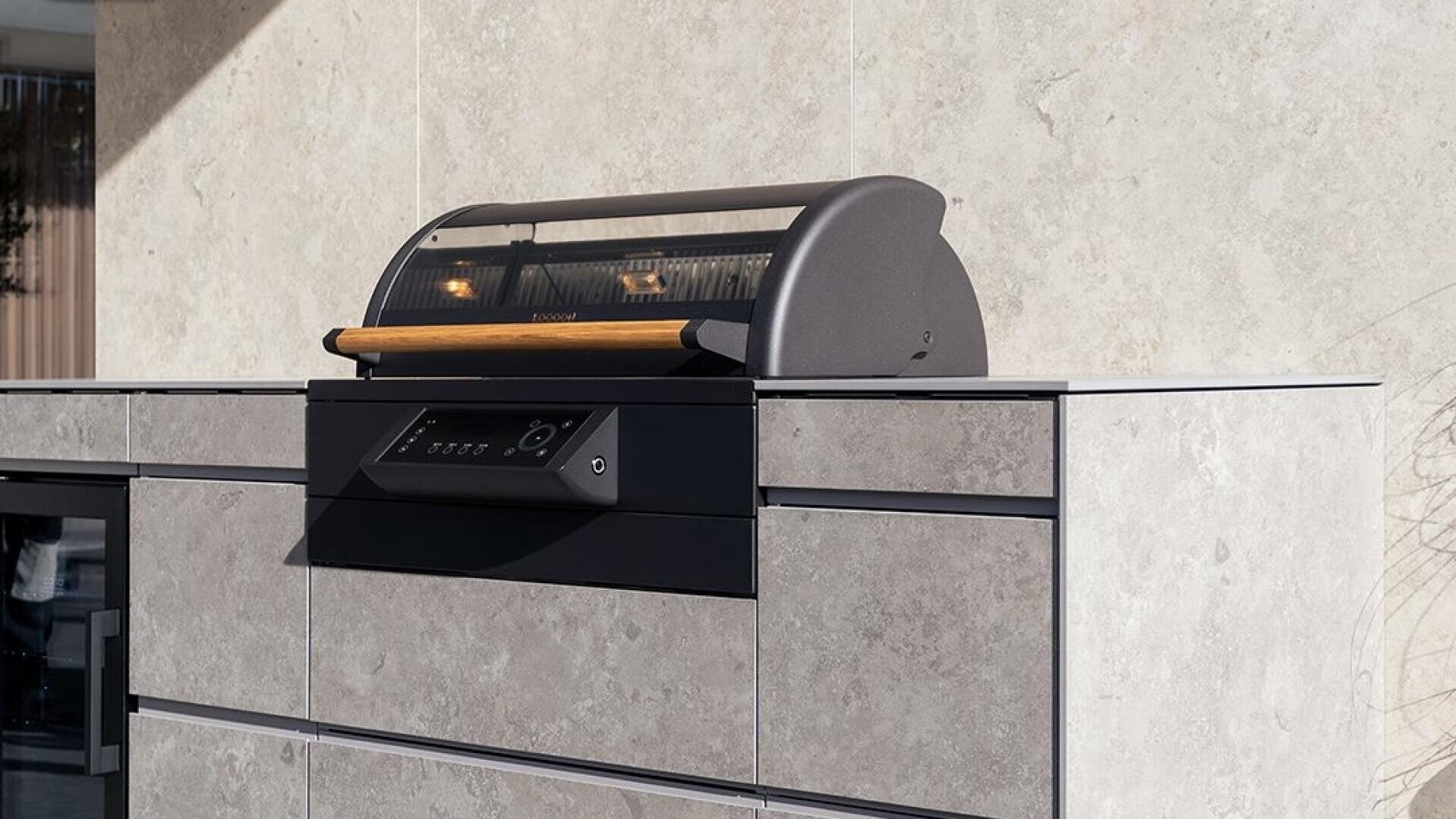 A modern outdoor grill with a curved lid and wooden handle is built into a sleek, light gray stone countertop and cabinet set against a matching stone wall.