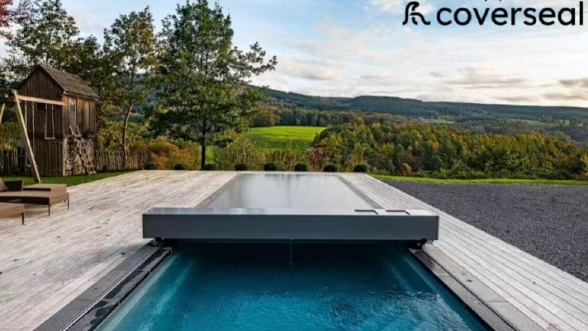 Eine elegante automatische Poolabdeckung schließt sich teilweise über einem modernen Außenpool auf einem Holzdeck mit Blick auf eine üppige grüne Landschaft mit Bäumen und sanften Hügeln unter einem teilweise bewölkten Himmel.