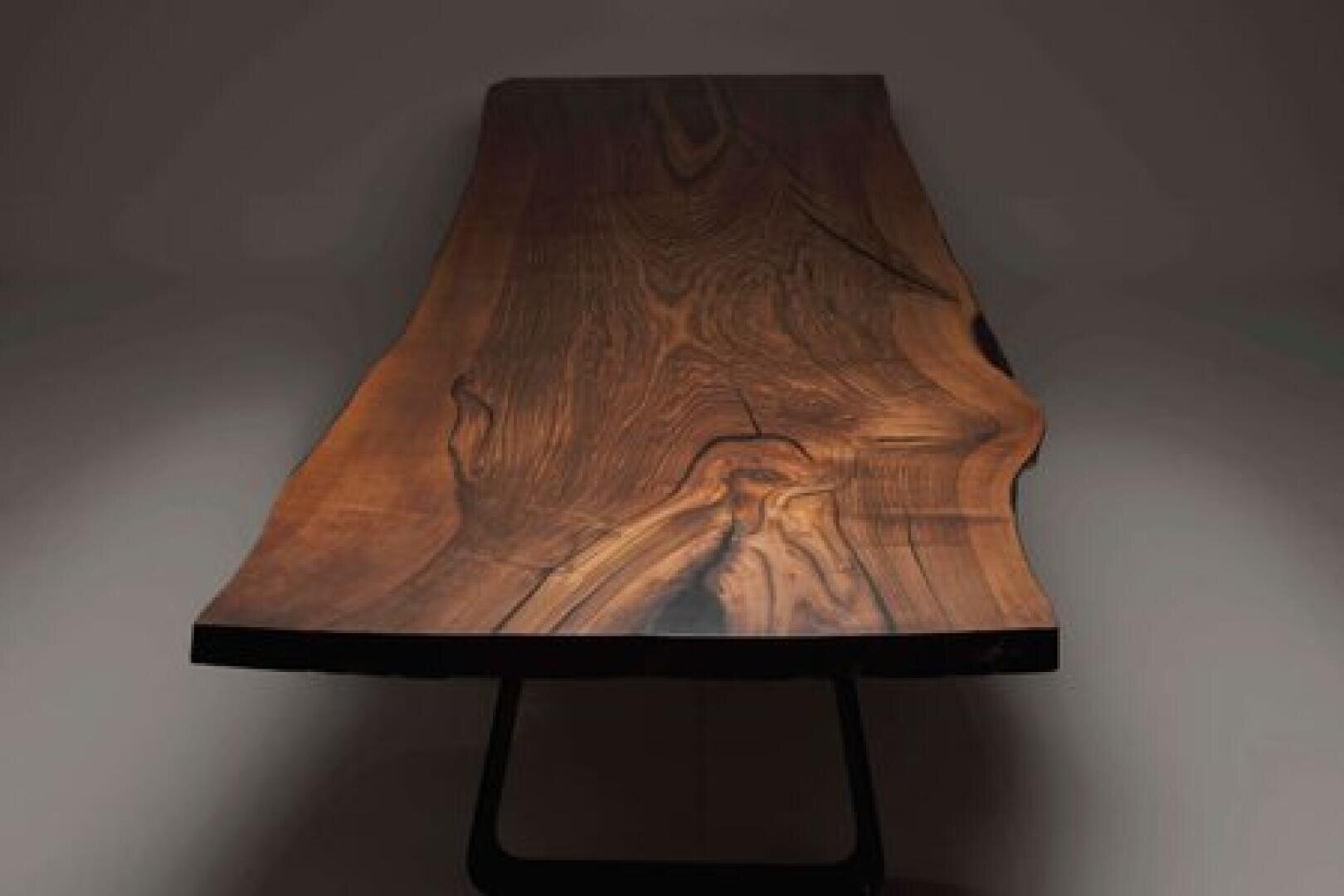 A close-up view of a polished wooden table with a natural, irregular edge and visible wood grain patterns, set against a plain, dimly lit background.