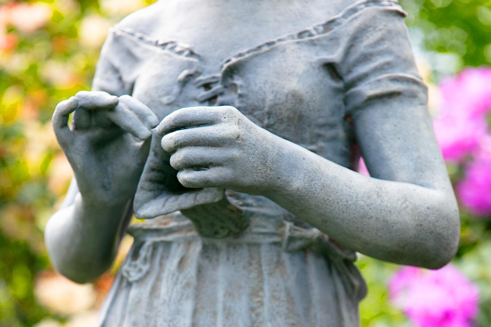 Nahaufnahme einer verwitterten Steinstatue eines Mädchens, wobei der Schwerpunkt auf ihrem Oberkörper und ihren Händen liegt, die einen kleinen Beutel halten. Der Hintergrund ist mit grünem Laub und rosa Blumen verschwommen.