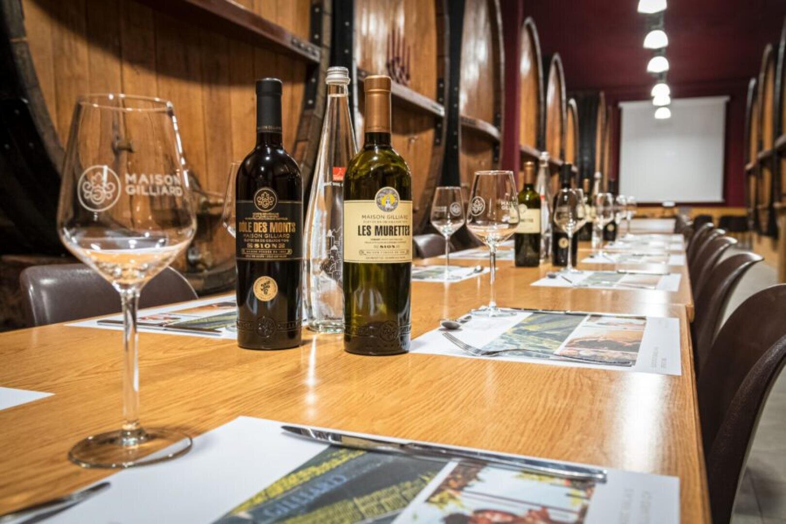 A long wooden table set for a wine tasting, with wine bottles, glasses, water bottles, and placemats arranged neatly, surrounded by wooden barrels in a warmly lit cellar.