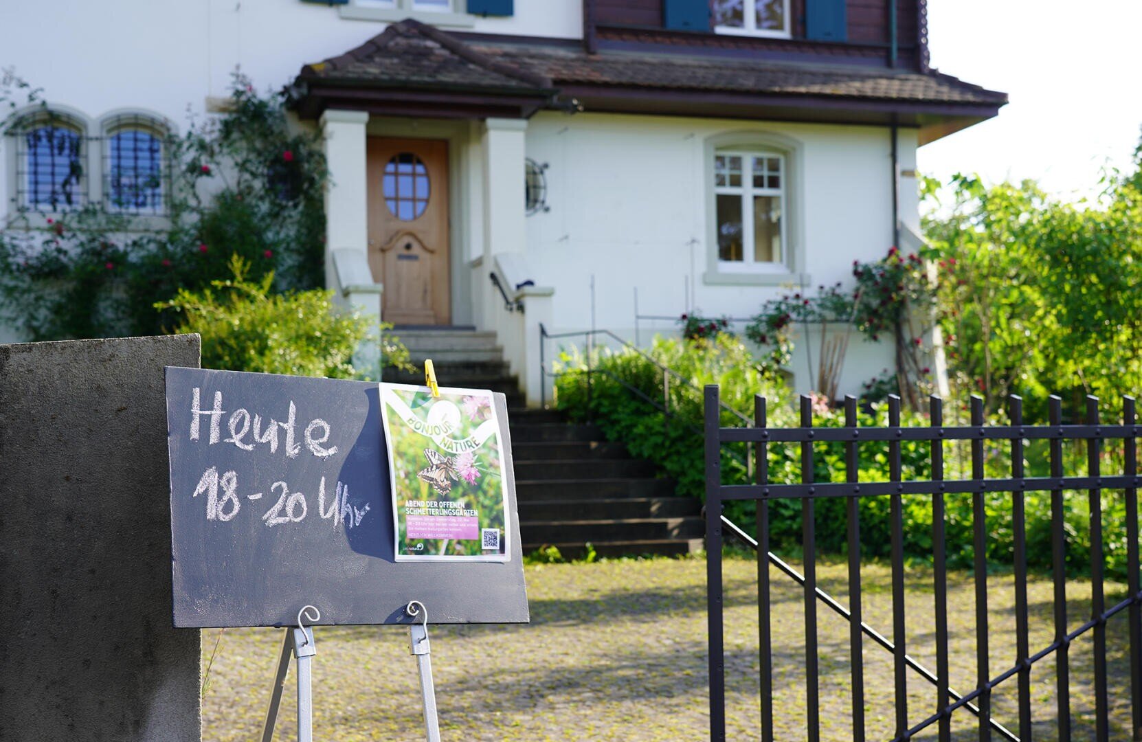 Auf einem Kreideschild vor einem Haus steht "Heute 18-20 Uhr", daneben ein Flugblatt. Das Haus hat eine Holztür, Rundbogenfenster und ist von Grün und Blumen umgeben. Im Vordergrund ist ein schwarzes Metalltor teilweise geöffnet.