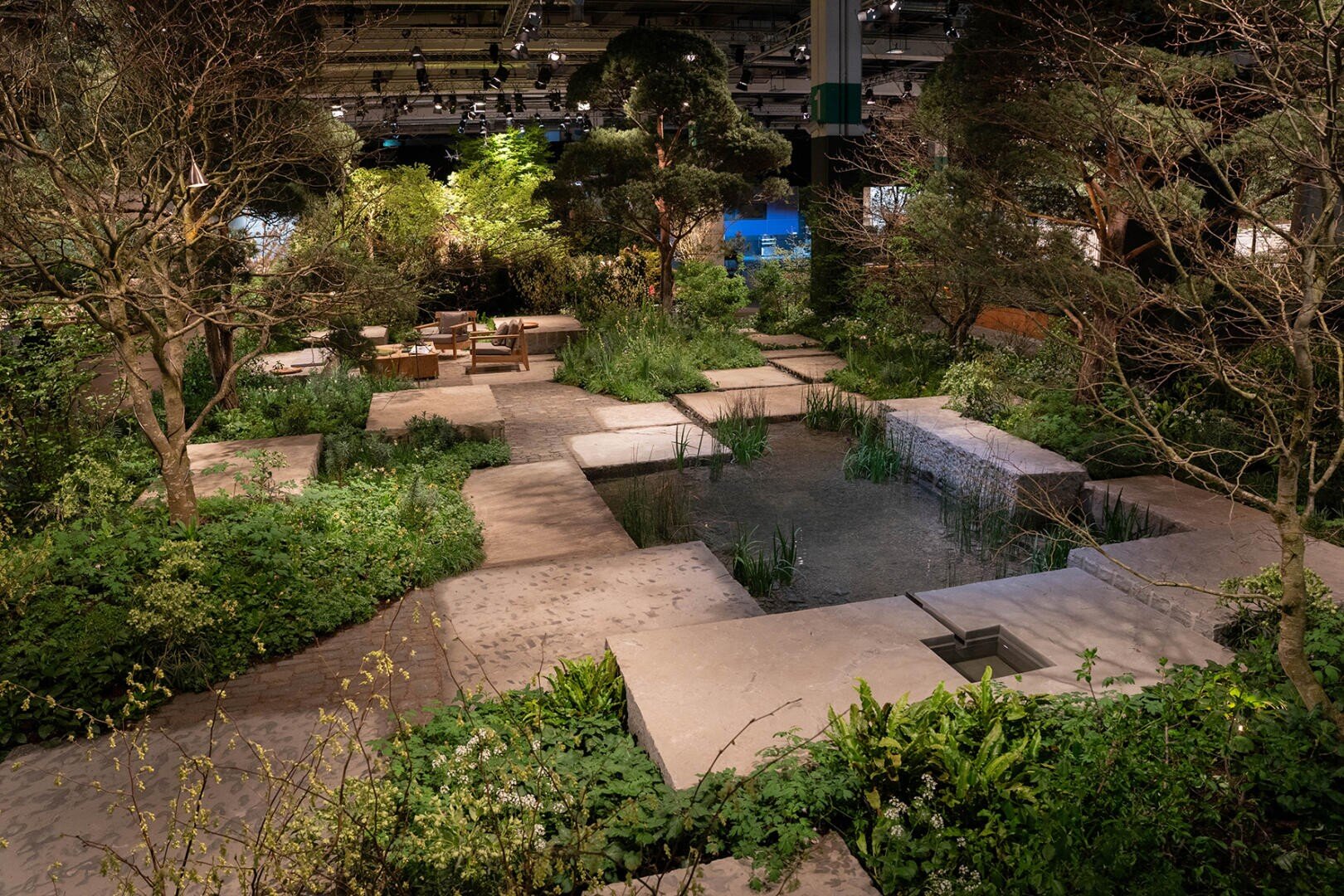 Moderner Innengarten mit einem zentralen Teich, umgeben von üppigem Grün, Bäumen und Steinwegen. Inmitten der Vegetation stehen Holztische und -stühle, die von einer sanften, warmen Beleuchtung umgeben sind.