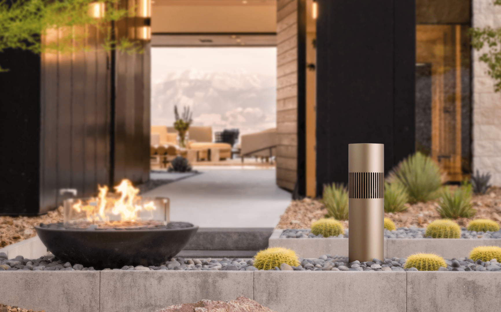 A modern outdoor terrace with a round fireplace, small cacti and rocks in the foreground. A simple, cylindrical metal lamp stands nearby, while blurred mountains and modern furniture can be seen in the background.