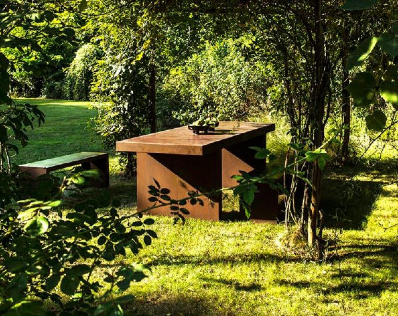 A wooden table and bench sit on grass under leafy trees in a shaded, sun-dappled garden. A bowl with fruit or plants is on the table, surrounded by lush greenery and natural light.