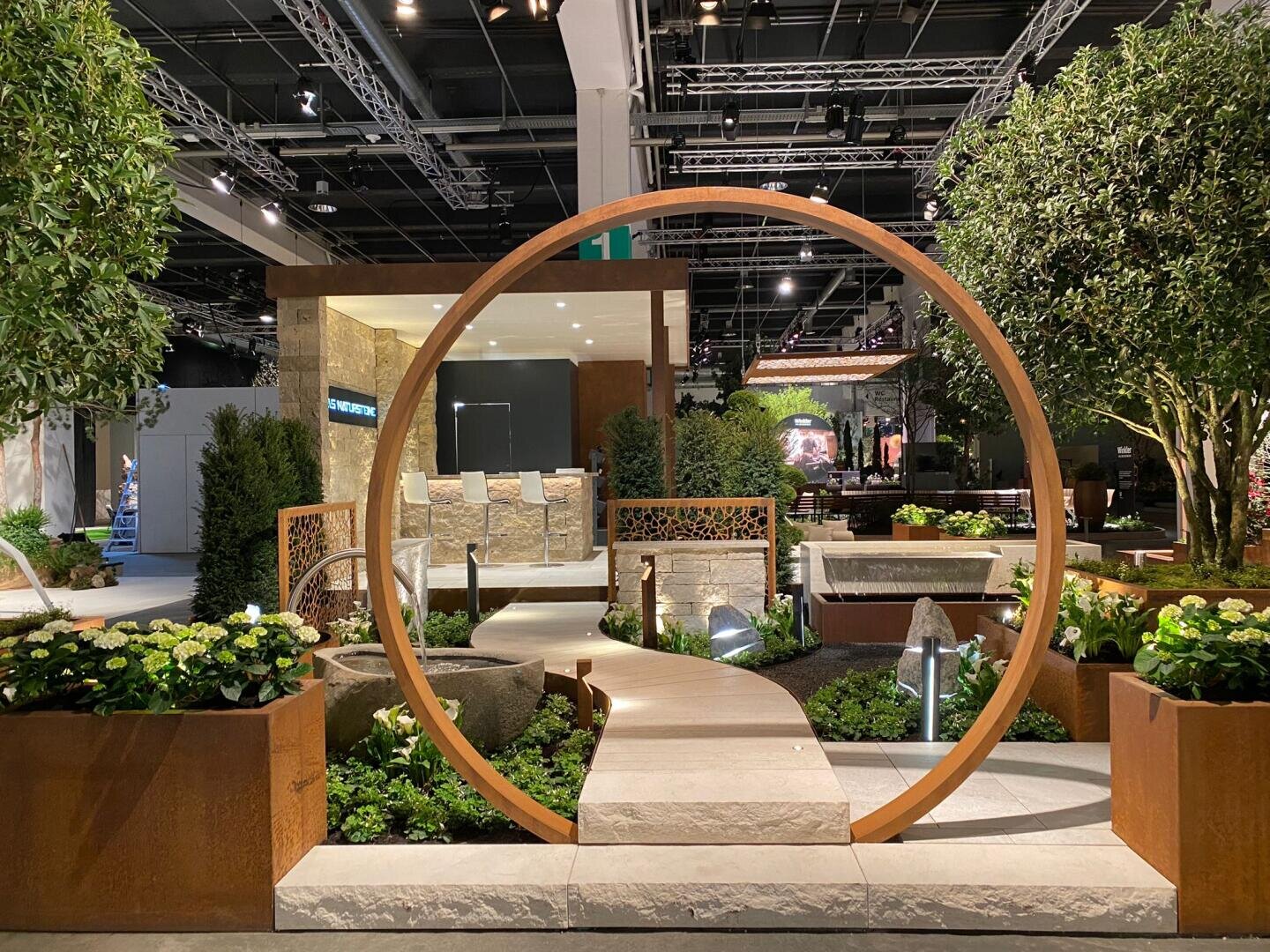 Ein moderner Indoor-Garten zeigt einen großen runden Metallbogen über einem Steinweg, umgeben von üppigem Grün, Blumen und Springbrunnen, mit Bäumen und architektonischen Elementen im Hintergrund.