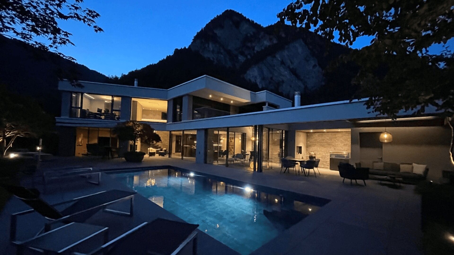 Modernes, luxuriöses Haus mit großen Glasfenstern und einem beleuchteten Swimmingpool in der Abenddämmerung, umgeben von Bergen und Bäumen. Die Terrassenmöbel sind um den Pool herum angeordnet, und die Innenbeleuchtung sorgt für ein warmes Licht.