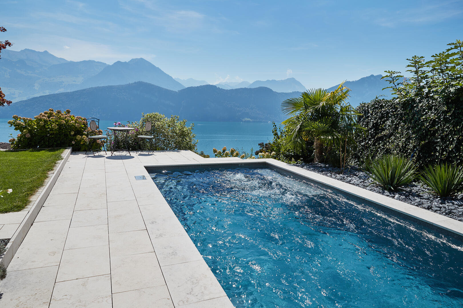 Ein rechteckiger Swimmingpool neben einer Steinterrasse bietet einen Blick auf einen großen blauen See und ferne Berge unter einem klaren Himmel, umgeben von üppigem Grün und Palmen.