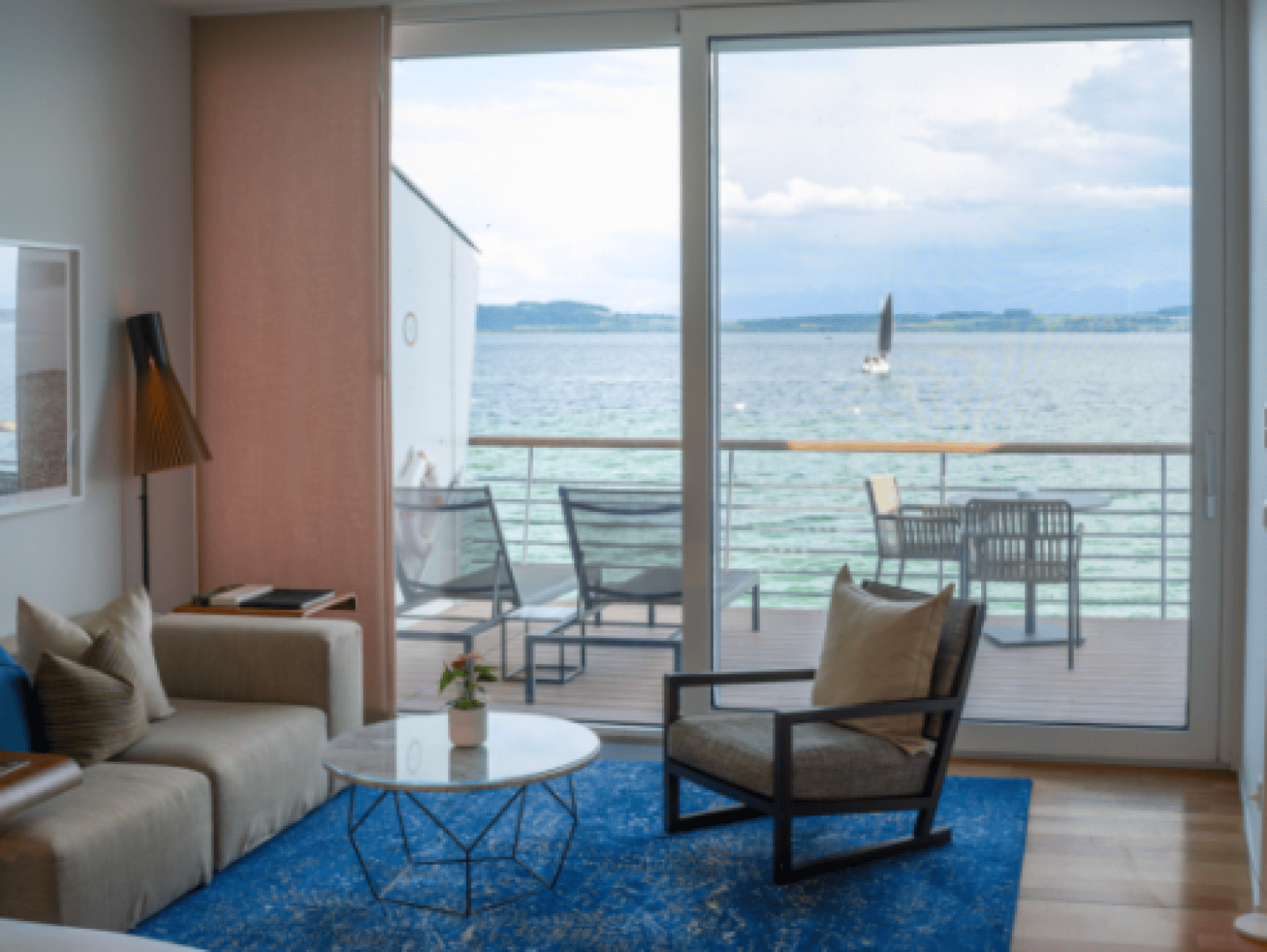 A cozy living room with sofa, armchair and round coffee table opens onto a balcony overlooking a tranquil lake, with a sailboat on the water and outdoor seating on the deck.