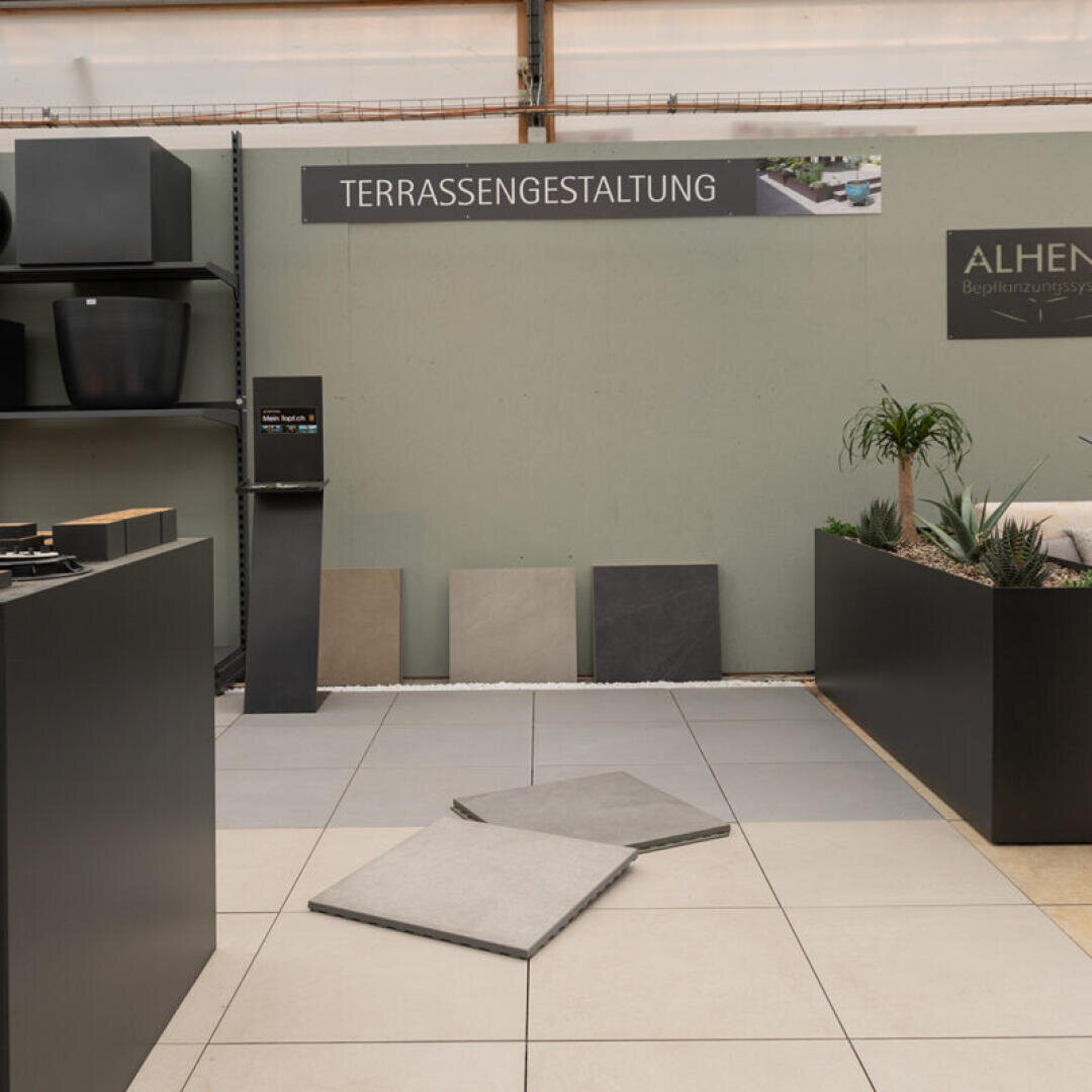 Ausstellungsbereich für Terrassengestaltung mit mehreren Mustern von Fliesen für den Außenbereich auf dem Boden und an der Wand, einem Schild mit der Aufschrift "Terrassengestaltung", Pflanzgefäßen auf Regalen und einem Pflanzgefäß mit Grünpflanzen auf der rechten Seite.