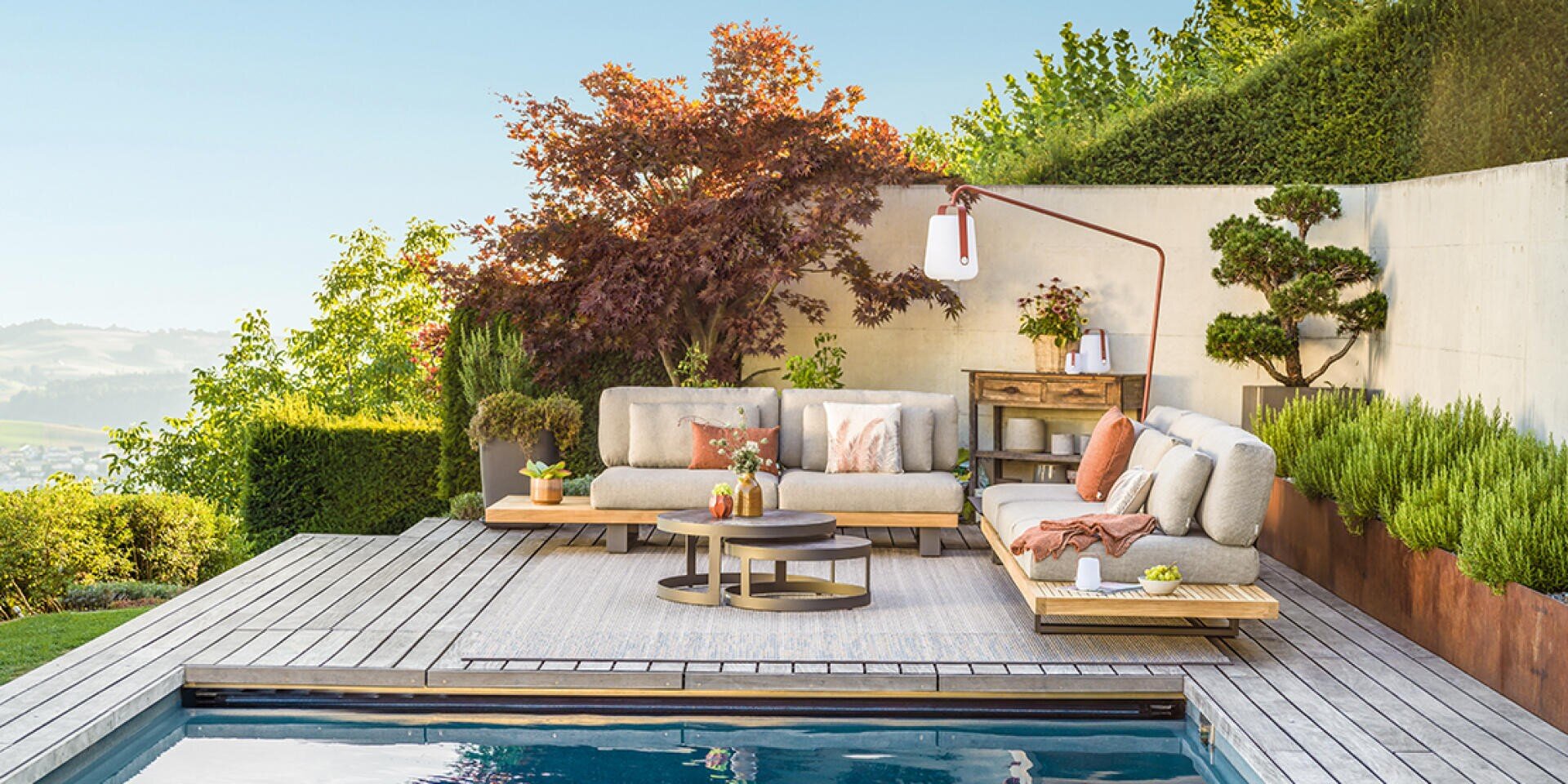 Moderne Außenterrasse mit hellgrauen Sofas, Couchtischen und Zierkissen neben einem Swimmingpool. Üppiges Grün und ein Baum rahmen den Platz ein, im Hintergrund sind unter dem klaren Himmel die Hügel zu sehen.