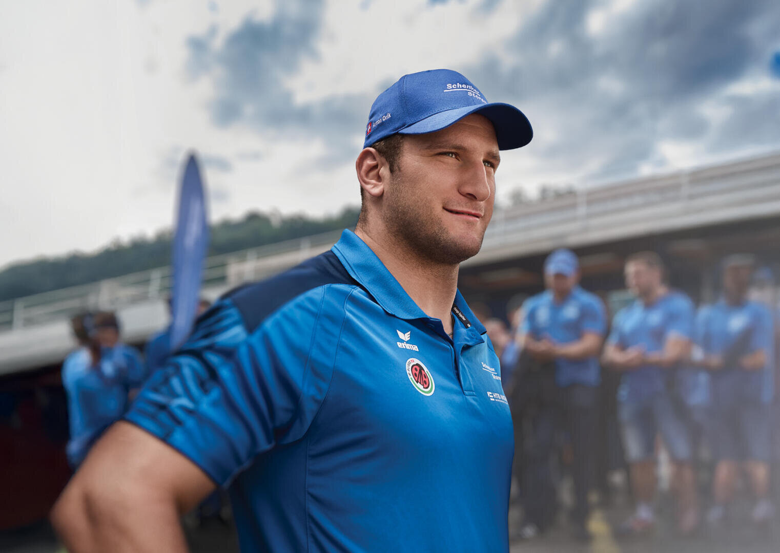 Ein Mann in einem blauen Sportdress und einer Mütze steht selbstbewusst im Freien, während eine Gruppe ähnlich gekleideter Menschen im Hintergrund verschwimmt. Der Himmel ist bewölkt, und hinter ihnen steht ein Gebäude.