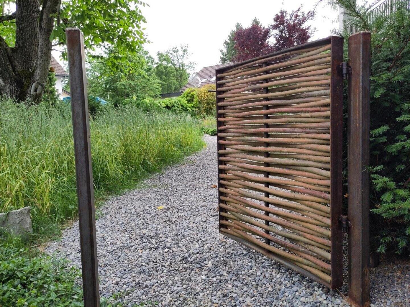 A wooden gate made of horizontal branches stands open and provides a view of a gravel path lined with tall grass and greenery in a garden landscape. Trees and shrubs can be seen in the background.