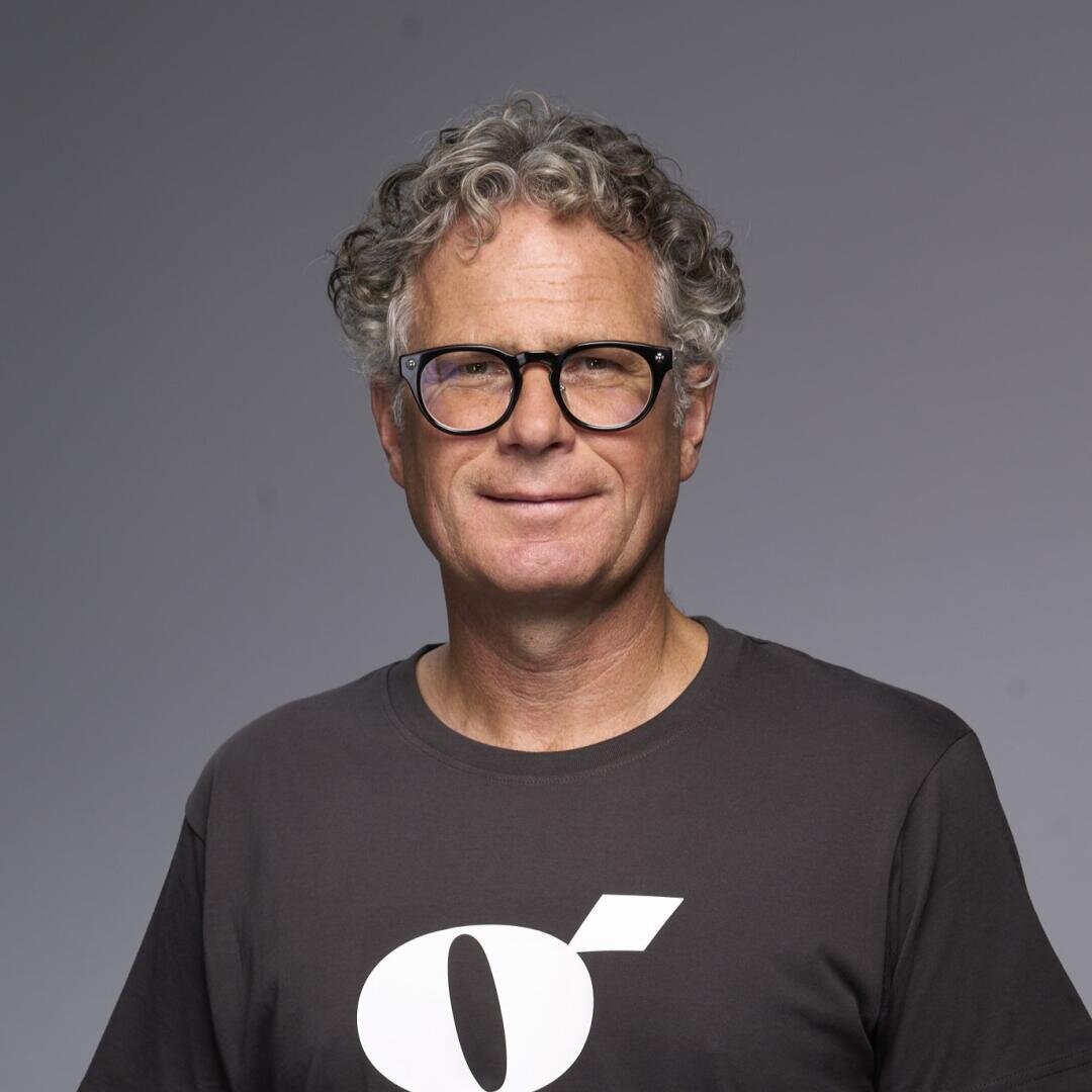 Ein Mann mittleren Alters mit lockigem grauem Haar und Brille, der ein dunkles T-Shirt mit einer auffälligen weißen Grafik trägt, steht vor einem einfarbigen grauen Hintergrund und lächelt leicht in die Kamera.